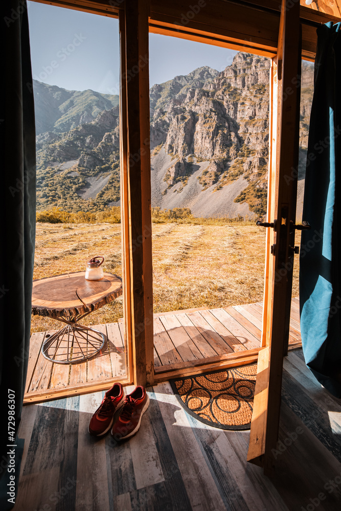 view from a small wooden chalet house in the mountains outside through a glass door and window. The concept of glamping and idyllic holidays