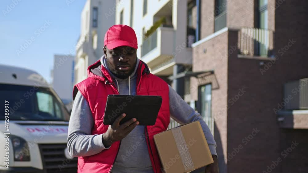 African american delivery man handling parcel box, walking along the street with tablet. Courier on the way to deliver postal parcel to a client. African american delivery man handling parcel box, walking along the street with tablet. Courier on the way to deliver postal parcel to a client.