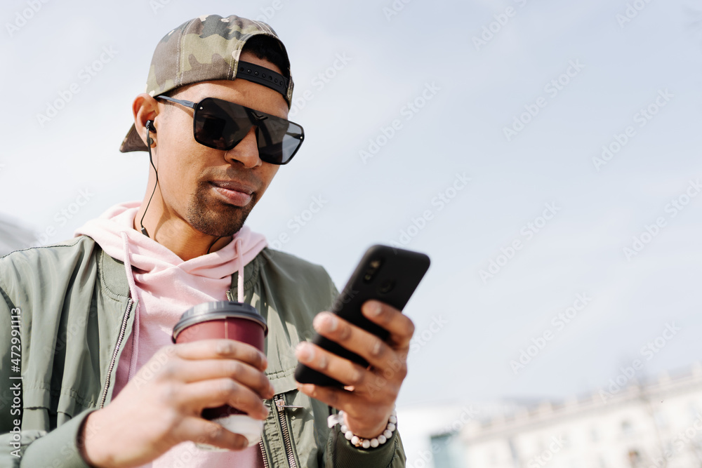People, technology, travel and tourism - Afro man using smartphone on city street bench and texting while drinking coffee People, technology, travel and tourism - Afro man using smartphone on city street bench and texting while drinking coffee