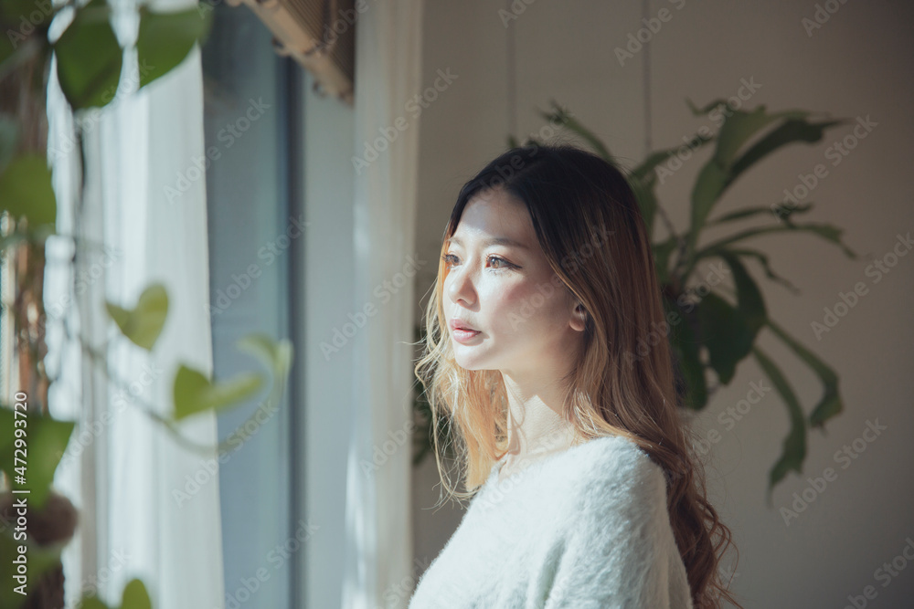 植物と光の差し込む窓辺に佇み、外を眺めるロングヘアの美しい女性 植物と光の差し込む窓辺に佇み、外を眺めるロングヘアの美しい女性
