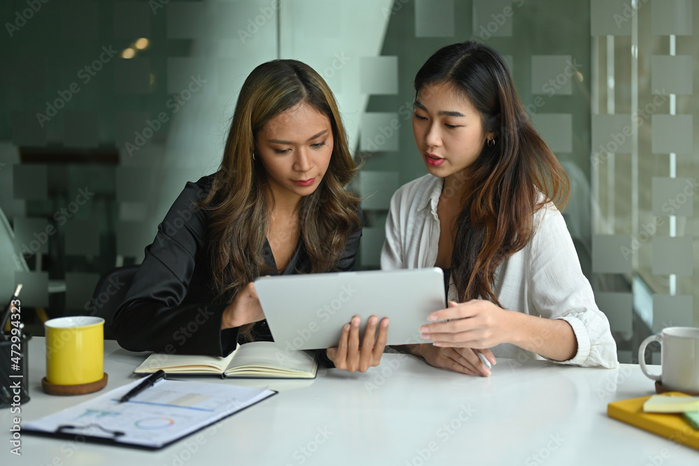 Photo of two female designers working together with a digital tablet at the white working desk in the comfortable living room. Photo of two female designers working together with a digital tablet at the white working desk in the comfortable living room.