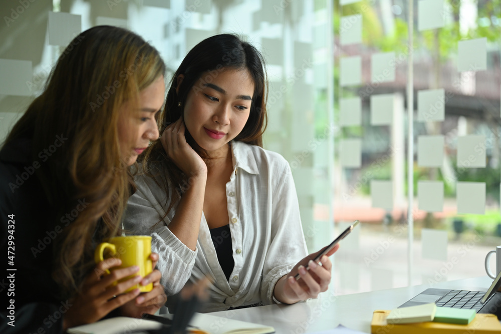 Photo of two office women relaxing with a smartphone in a coffee break time over the modern office as a background. Photo of two office women relaxing with a smartphone in a coffee break time over the modern office as a background.