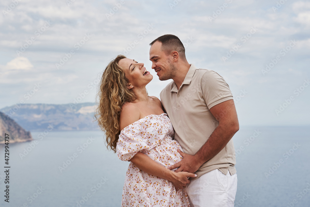 Fototapeta premium happy and very beautiful young couple expecting a baby. a pregnant woman laughs, and a happy man holds her belly against the backdrop of a beautiful seascape