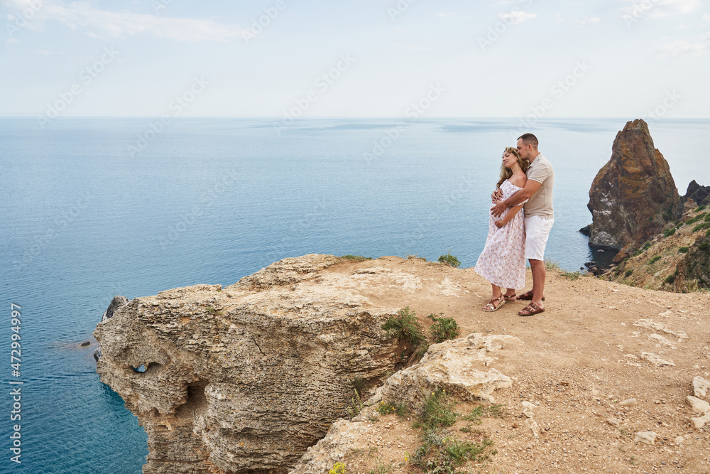 a man and a pregnant woman against the backdrop of the sea receding into the distance. a man hugs a woman from the back. family, travel, vacation, beautiful landscape, love