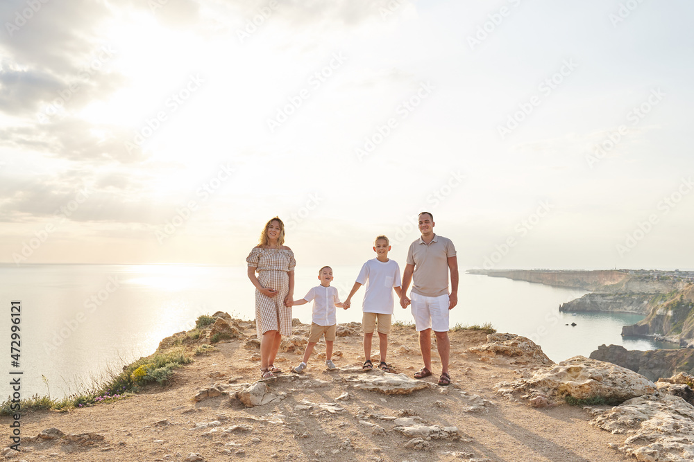 a big family against the backdrop of a beautiful warm landscape. mom, dad and two sons are all holding hands. mom is pregnant