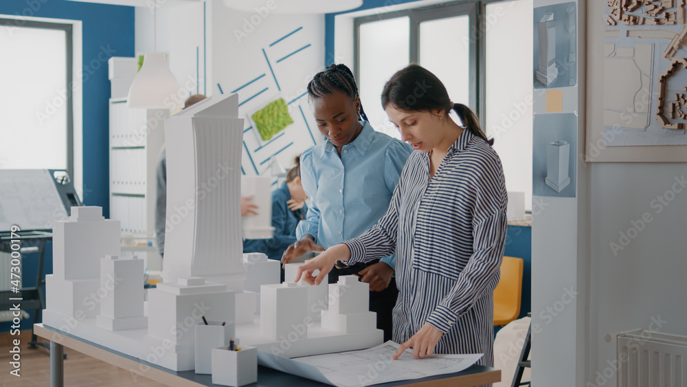 Multi ethnic women analyzing building model and blueprints plan to develop construction. Architectural coworkers brainstorming ideas and working on property layout with maquette and sketch. Multi ethnic women analyzing building model and blueprints plan to develop construction. Architectural coworkers brainstorming ideas and working on property layout with maquette and sketch.