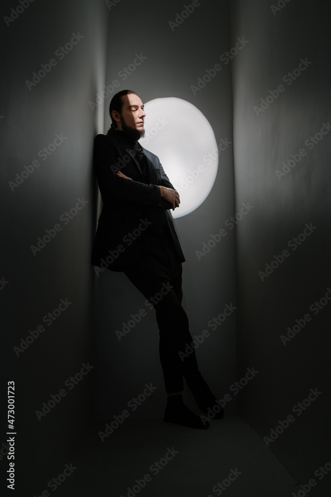 Fototapeta premium Calm sad handsome stylish man with long hair and beard in suit leans diagonally against the wall and looks thoughtfully down. Fashion style posing. Dark colors vertical composition with spotlight