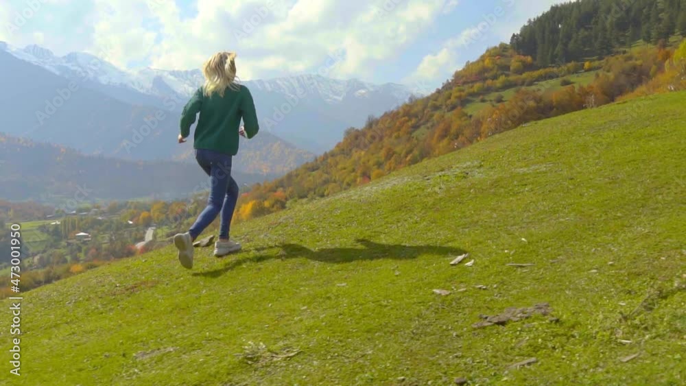 camera follows young beautiful woman who runs uphill on slope against background of valley of mountains. Autumn. runs all happy and joyful, inspired by landscape. Blonde in green sweater And jeans. camera follows young beautiful woman who runs uphill on slope against background of valley of mountains. Autumn. runs all happy and joyful, inspired by landscape. Blonde in green sweater And jeans.