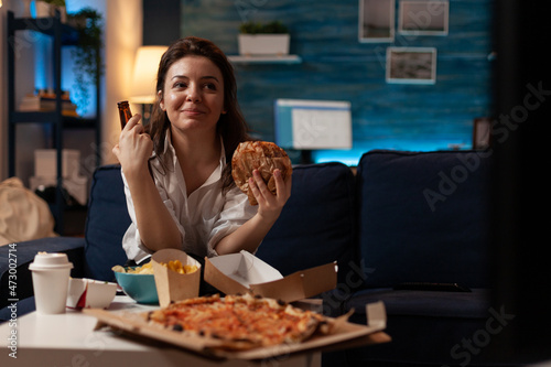 Woman eating tasty takeaway fast food hamburger while enjoying a cold beer in front of television with comedy show in home living room. Smiling person having hot pizza and takeout delivery menu.