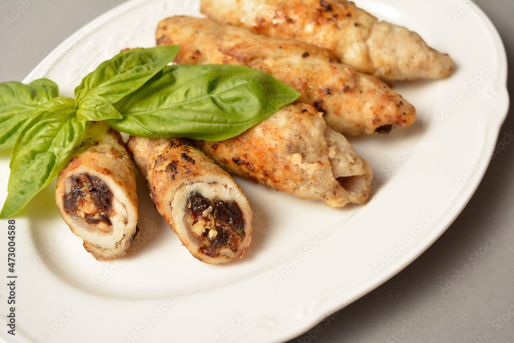 Meat roll with dried apricots, prunes and walnuts inside. Chicken Meat cannelloni stuffed with dried fruits and nuts. Served with fresh basil leaf on white vintage plate on gray solid background 
