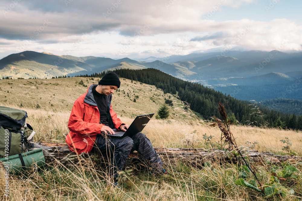 Caucasian male hiker working with laptop sitting on the mountain against the backdrop of beautiful scenic rocks.