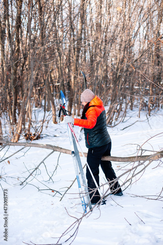 Young woman is doing sports in forest. Brunette in winter tracksuit walks along snowy road and holds ski poles and skis in her hands, side view. 