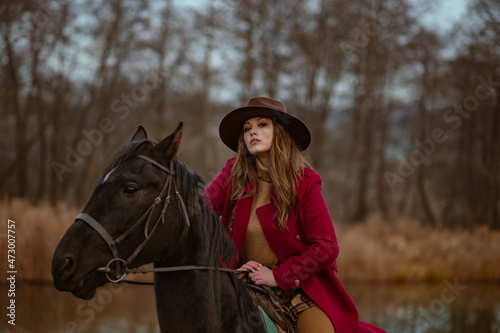 Elegant fashionable confident woman wearing trendy brown hat, classic red marsala color woolen coat posing, riding a horse in nature. Outdoor autumn fashion portrait. Copy, empty space for text 