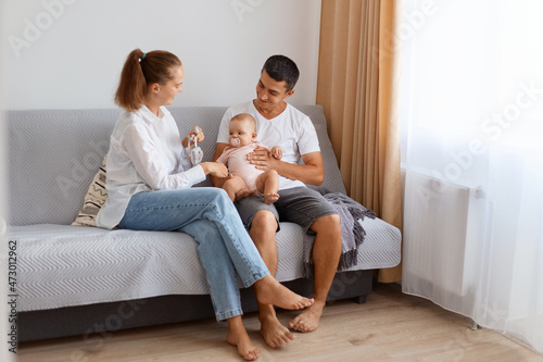 Romantic satisfied young couple wearing casual style clothing spending time together with their child, sitting on sofa in light living room with infant baby, parenthood.