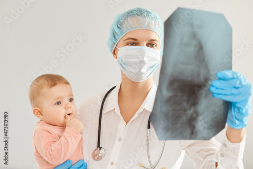 Portrait of young adult female pediatrist wearing medical uniform, holding infant baby girl, examining child's health in clinic, looking at x ray, trying to diagnose the kid correctly.