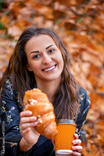 Smiling girl eating croissant and coffee in autumn park sitting on blanket