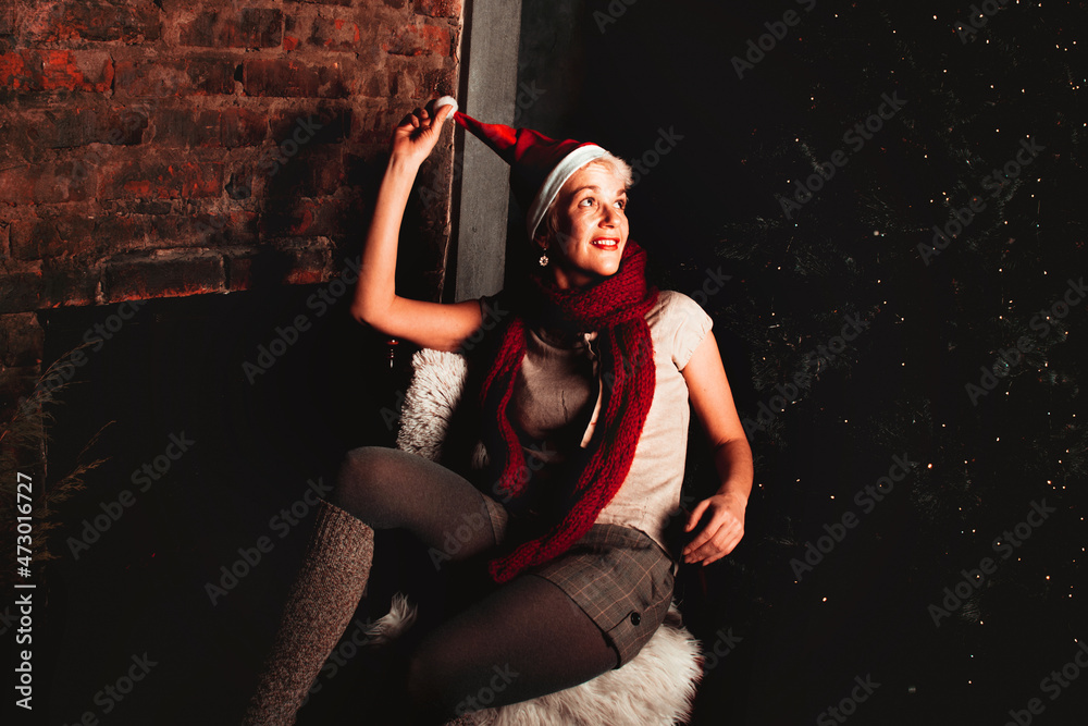 Beautiful middle-aged woman wearing a stocking cap or Christmas Santa Claus cap sitting in chair at New Year tree Beautiful middle-aged woman wearing a stocking cap or Christmas Santa Claus cap sitting in chair at New Year tree