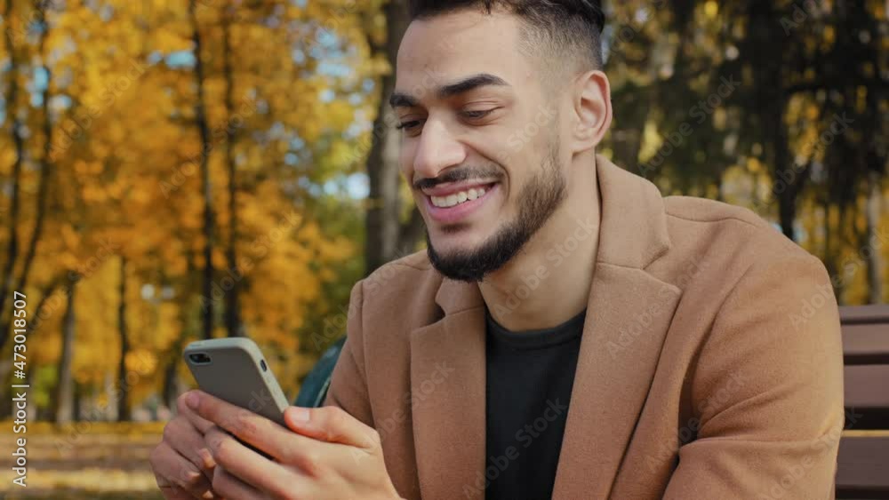Close up young hispanic guy sitting on bench in autumn park bearded man holding mobile phone makes online shopping smiling happily using app looking into smartphone outdoors browsing news on device