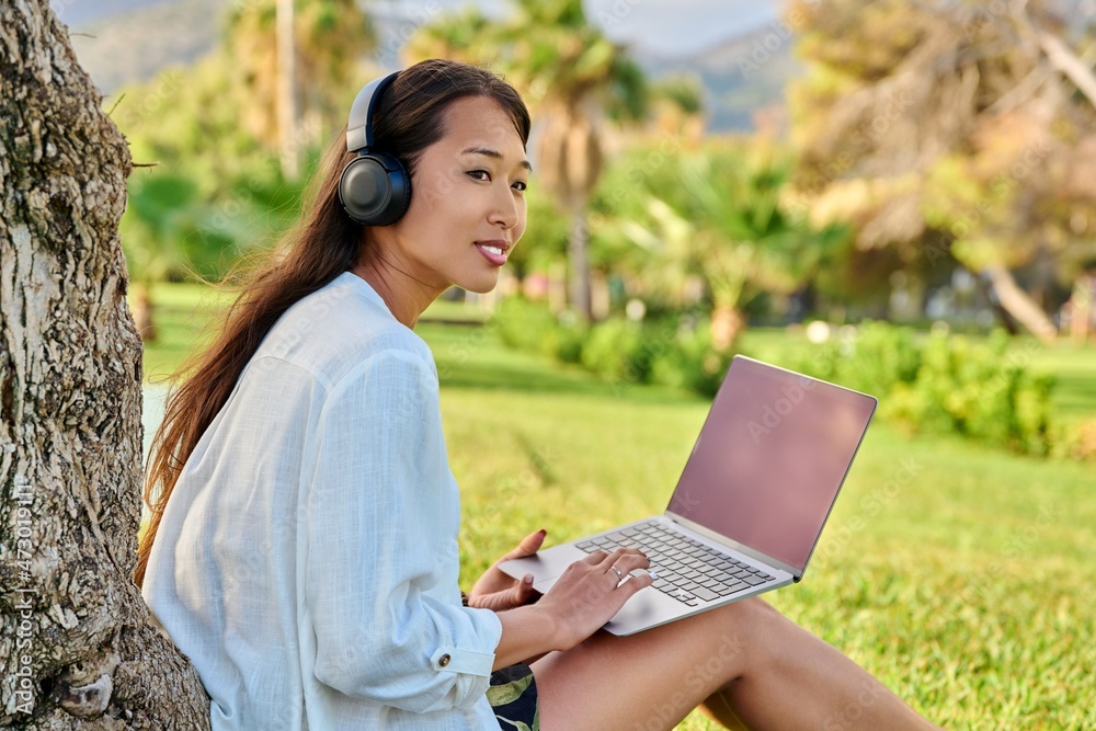 Young smiling woman in headphones using laptop sitting in park on grass Young smiling woman in headphones using laptop sitting in park on grass