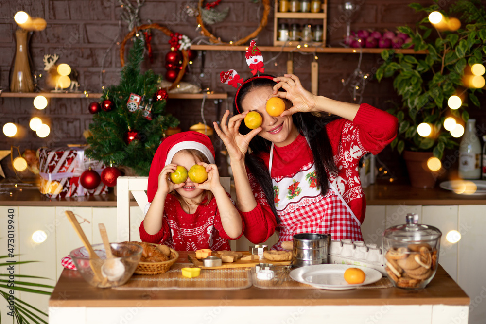 mom and daughter in a dark kitchen with a Christmas tree are preparing ginger cookies for New Year or Christmas, smiling and fooling around with tangerines in a Santa Claus hat