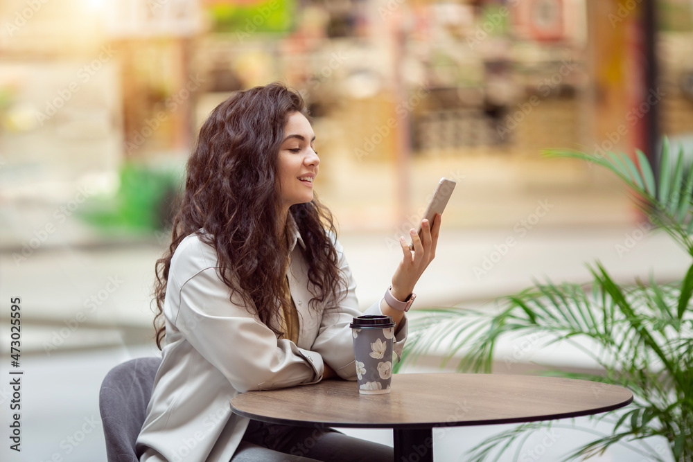 Young cheerfful woman using smartphone and drinking take away coffee in paper cup. Stylish woman sitting at cafe indoors.