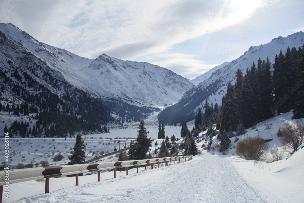 In winter, a snow-covered alpine gorge Alatau with the Big Almaty Lake, a mountain asphalt road in the snow, Christmas trees grow on the mountain slopes, a sky with clouds, sunlight