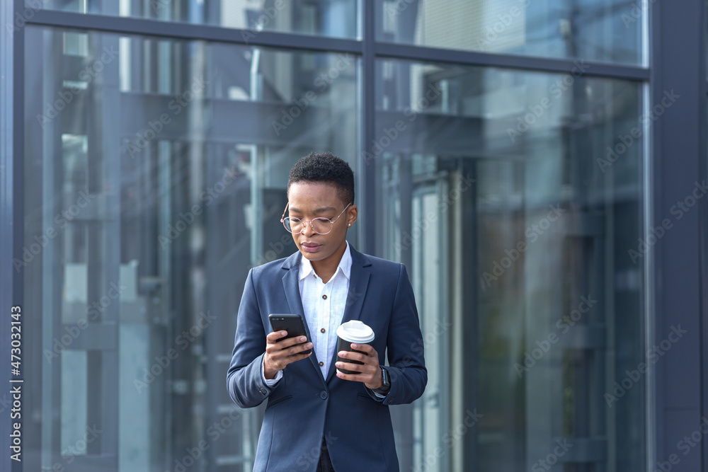 A confident African-American lady walks around the business district and uses the phone. Holding a cup of coffee, dressed in formal clothes. Attractive businesswoman browsing modern smartphone A confident African-American lady walks around the business district and uses the phone. Holding a cup of coffee, dressed in formal clothes. Attractive businesswoman browsing modern smartphone