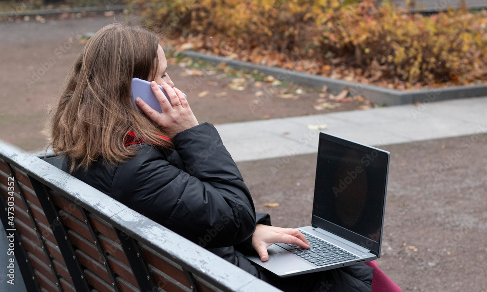 girl works on the computer while sitting on a park bench and speaking by the mobile phone girl works on the computer while sitting on a park bench and speaking by the mobile phone