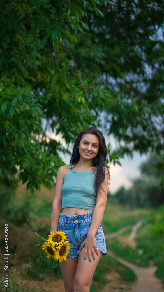 Portrait of a woman with sunflowers. The girl walks in the park with flowers. She is wearing denim shorts and a top. Leisure . Portrait of a woman with sunflowers. The girl walks in the park with flowers. She is wearing denim shorts and a top. Leisure .