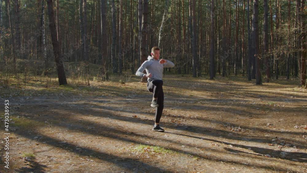 Young man is training outdoor, doing basic squats in combination with kicks, enjoying his intense cardio in forest, active morning time, Zoom out, Slow motion. Young man is training outdoor, doing basic squats in combination with kicks, enjoying his intense cardio in forest, active morning time, Zoom out, Slow motion.