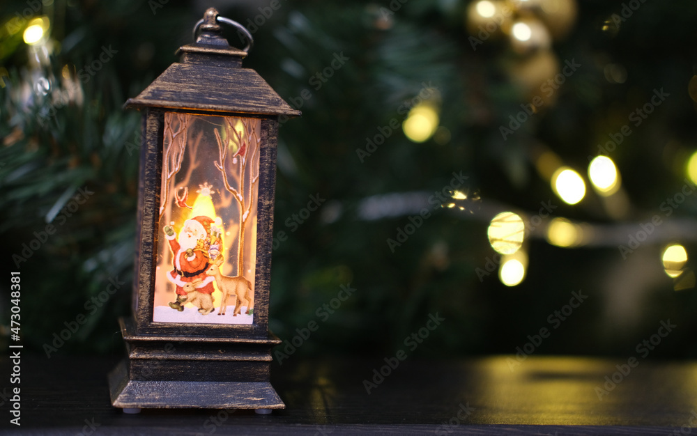 A lantern with candles inside stands against the background of a Christmas tree.