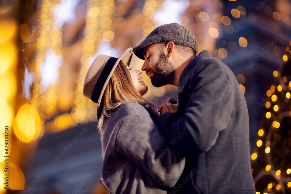Christmas couple kissing outdoors on New Year's eve. A young happy romantic couple standing on the street surrounded by Christmas lights, holding hands and kissing on New Year's eve.