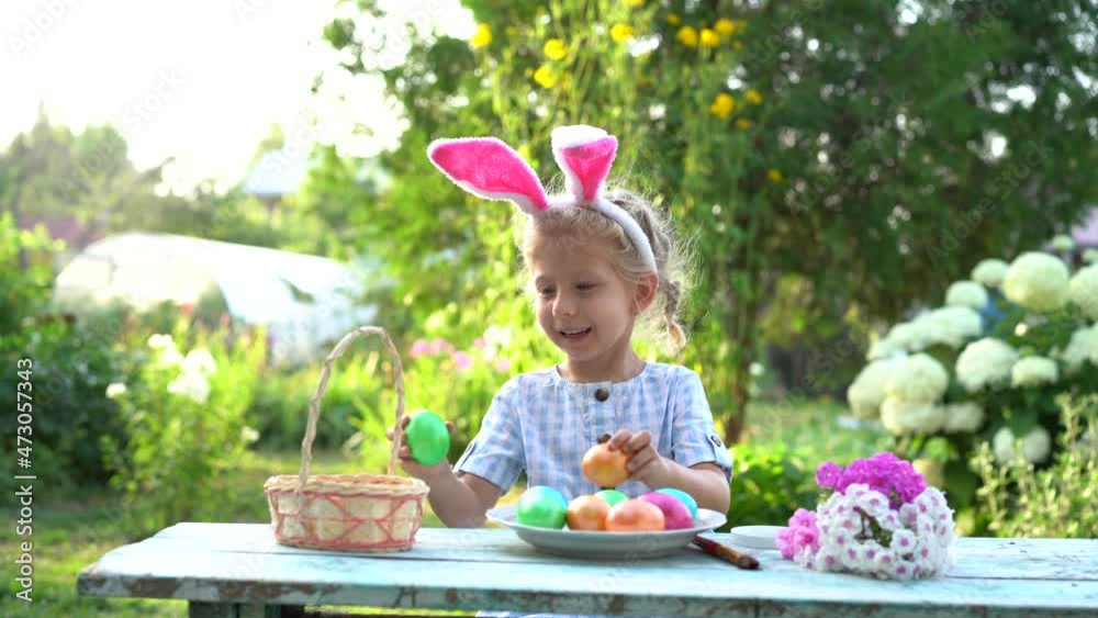 a little blonde girl with bunny ears puts colored Easter eggs in a basket in nature and smiles a little blonde girl with bunny ears puts colored Easter eggs in a basket in nature and smiles