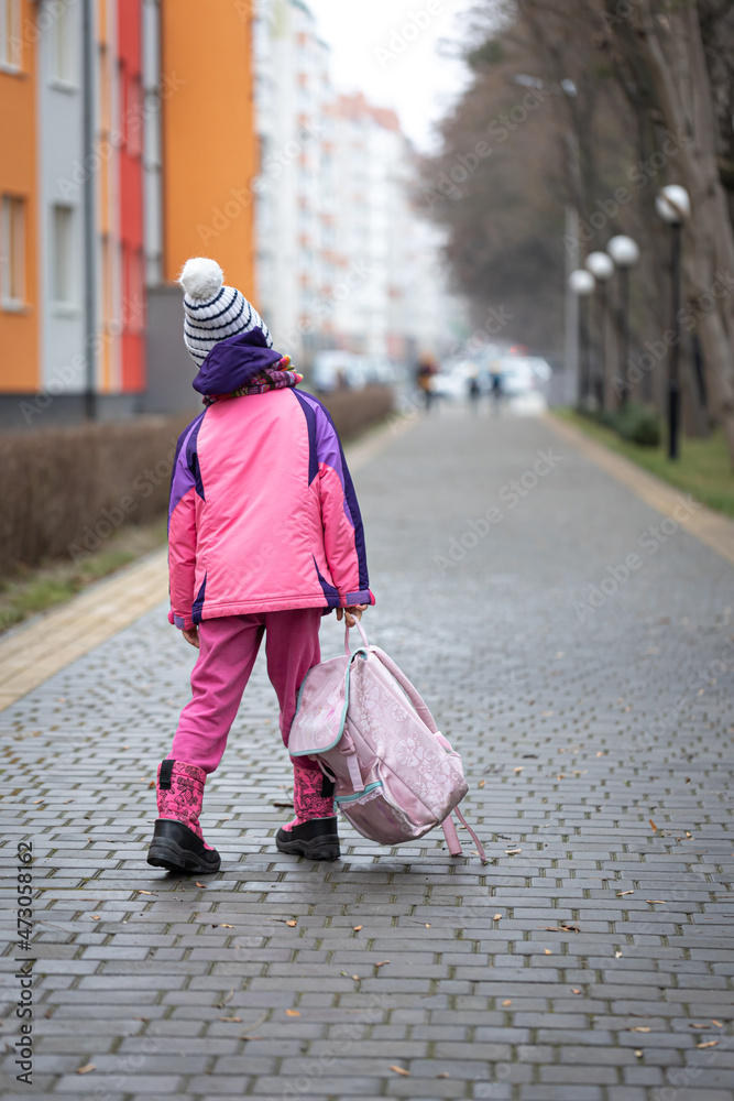 Fototapeta premium Little girl with a backpack, in a jacket and a hat near the school.