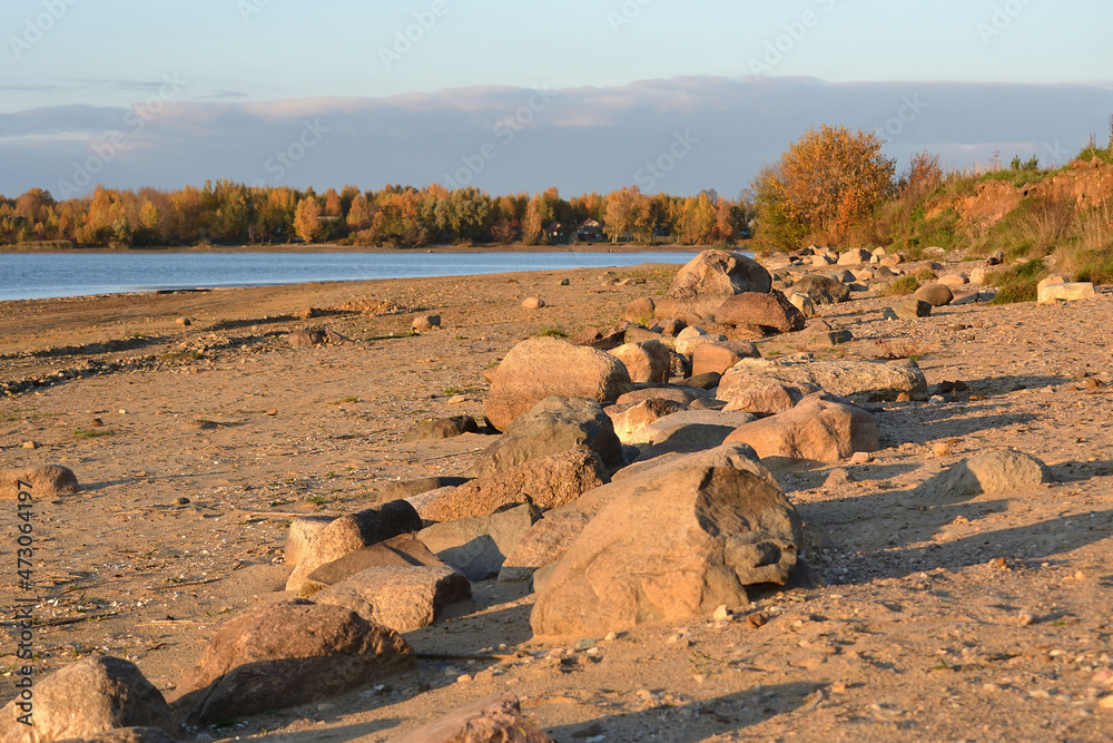 The drying bottom of the river against the background of a natural landscape - in the foreground there are large stones, boulders, sand, in the distance water, shore, trees, sky.