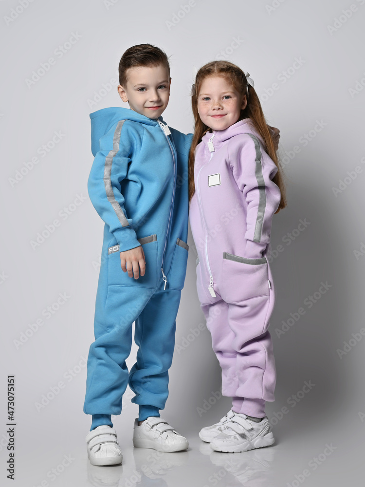 Tow smiling kids boy and girl, friends in blue and pink jumpsuits with hoods and pockets stand close together, looking at camera over light background