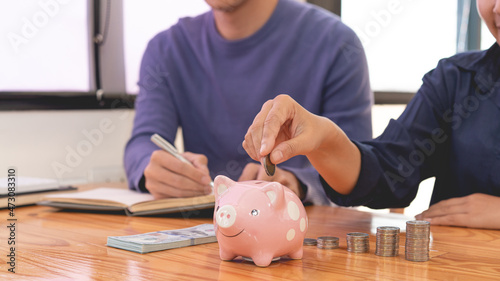 saving concept the man in blue sweater writing income account while the female in dark blue shirt drop a coin into a piggy bank