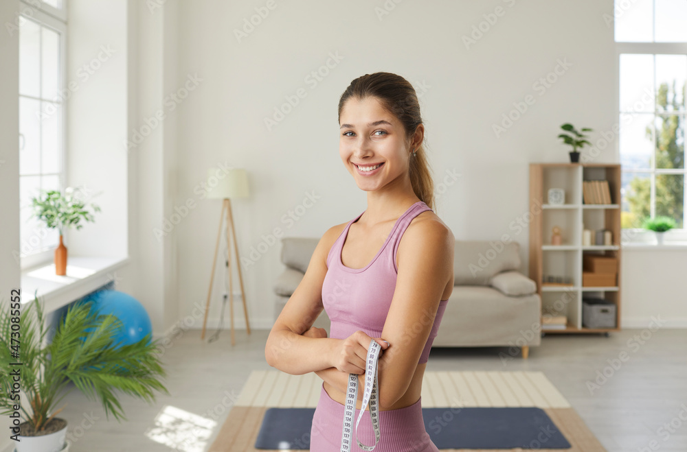 Portrait of happy slim woman with measuring tape. Cheerful attractive sportive young girl in sports bra top standing at home, holding tape measure, looking at camera and smiling. Weight loss concept Portrait of happy slim woman with measuring tape. Cheerful attractive sportive young girl in sports bra top standing at home, holding tape measure, looking at camera and smiling. Weight loss concept