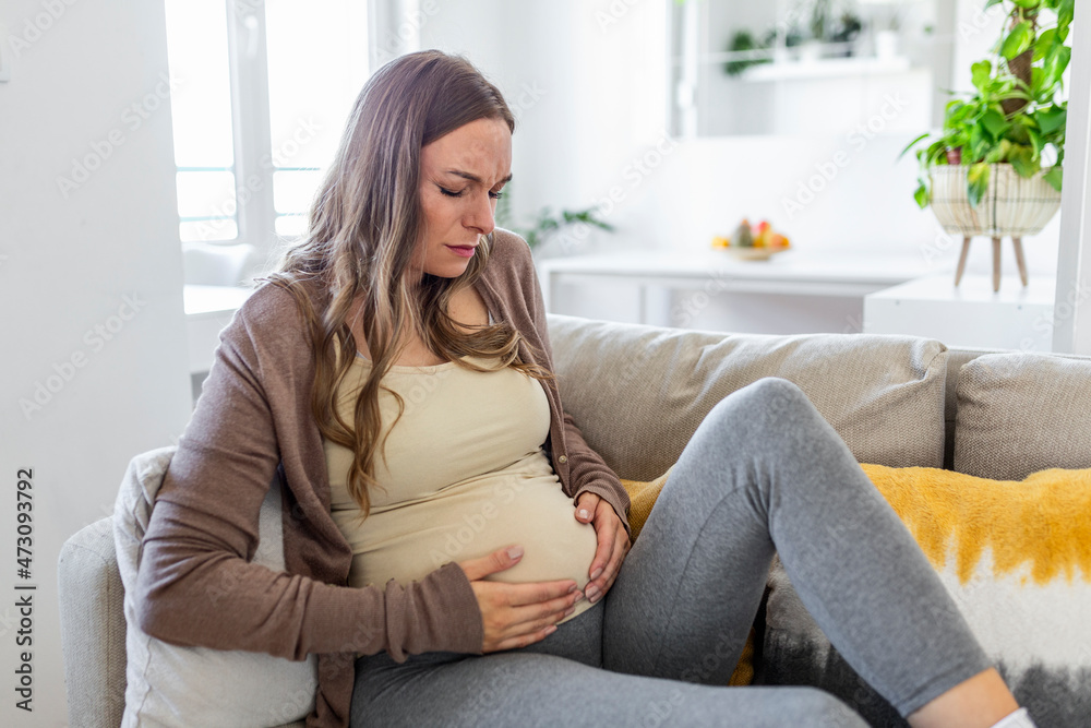 Pregnant woman suffering during her pregnancy, with back pain and headaches. Pregnant woman sitting on the sofa holding her belly with worried face expression. Pregnant woman suffering during her pregnancy, with back pain and headaches. Pregnant woman sitting on the sofa holding her belly with worried face expression.