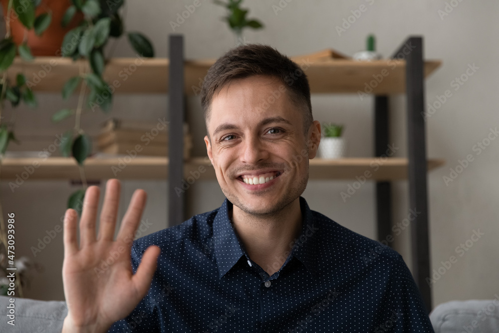 Naklejka premium Happy millennial business man in casual making video call from home couch, looking at camera, smiling, waving hand hello, attending online chat, virtual conference. Video call head shot portrait