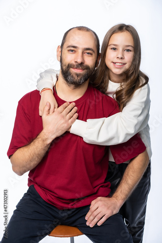 daughter and dad are looking at us in the studio. the child hugs the father. a man in a burgundy-red T-shirt, a girl in a white sweatshirt. isolated white background