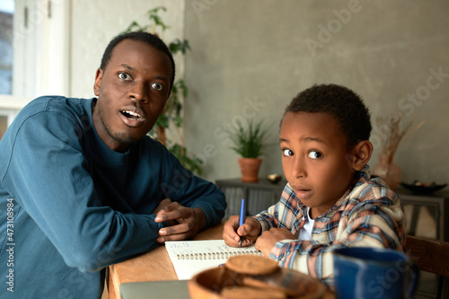 Indoor picture of dark-skinned male father sitting with his son at desk in living-room doing homework, having shocked face expressions, scared of exam failure, plate with cookies and mug on table