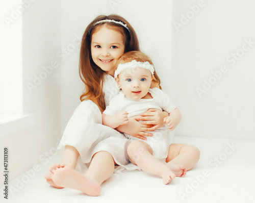 Two cute older and younger sisters children hugging together at home in white room near window