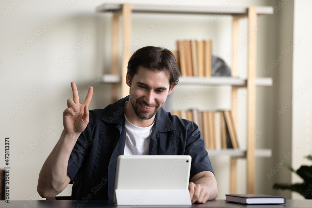 Positive millennial employee guy, tablet user waving hello at screen, showing peace, victory hand finger gesture, making distant video call to friend, coworker, client at workplace table Positive millennial employee guy, tablet user waving hello at screen, showing peace, victory hand finger gesture, making distant video call to friend, coworker, client at workplace table