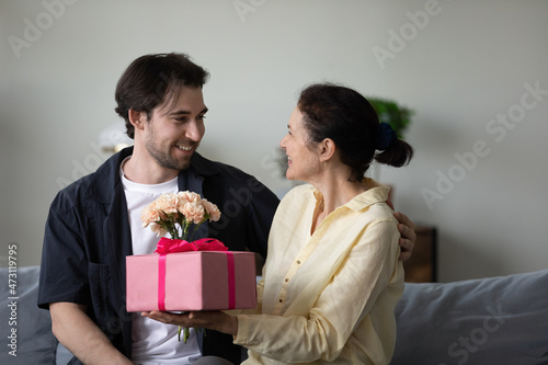 Excited cheerful middle aged mom receiving surprise gift and flower bunch from happy grateful adult son, celebrating birthday, mothers day, 8 march at home, holding present box, bouquet
