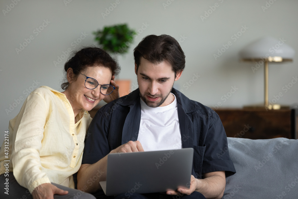 Adult son teaching mature mom to use online virtual app, banking service on laptop. Senior mother and grown child sitting on couch together, holding computer, browsing internet, talking Adult son teaching mature mom to use online virtual app, banking service on laptop. Senior mother and grown child sitting on couch together, holding computer, browsing internet, talking