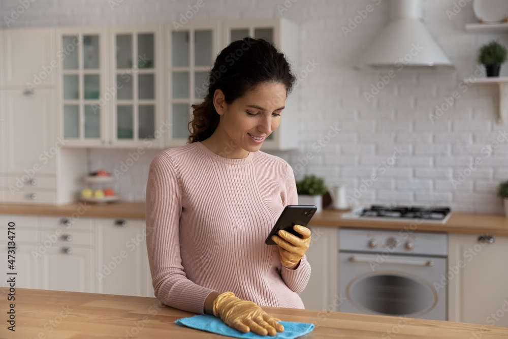 Smiling woman housewife using smartphone, distracted from cleaning, chatting online during break, standing in modern kitchen at home, smiling young female in gloves removing dust from wooden table
