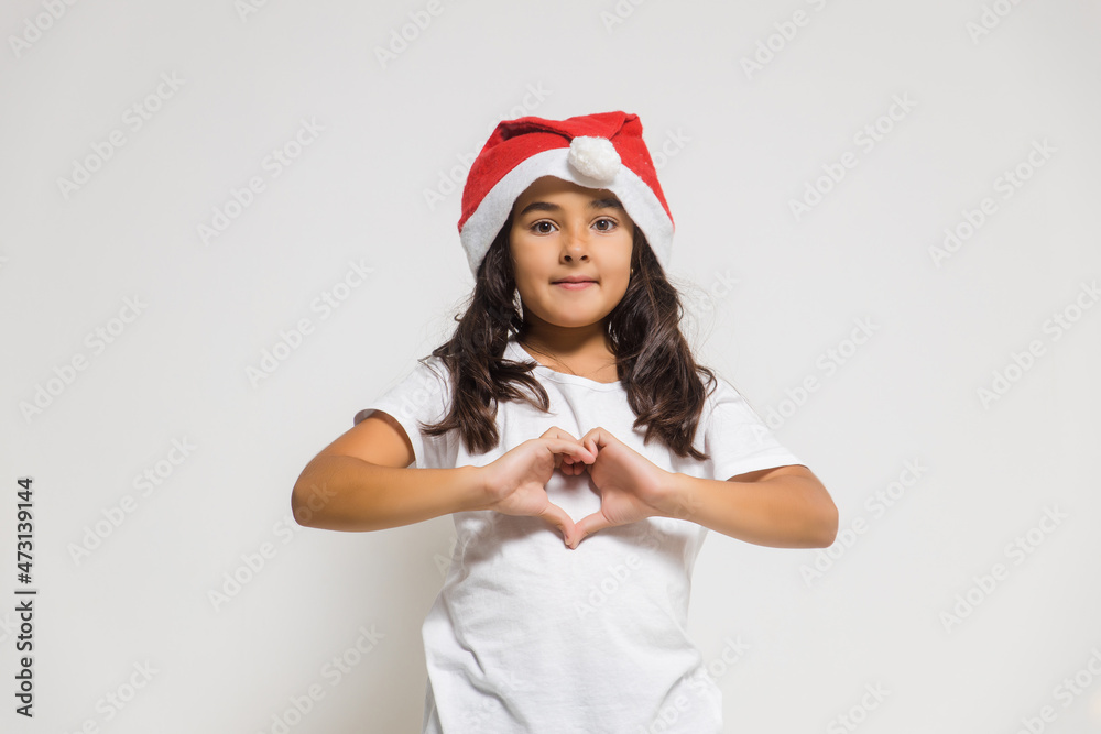 Smiling child girl in christmas santa hat showing heart gesture, congratulating with xmas holiday. Love, care and trust Smiling child girl in christmas santa hat showing heart gesture, congratulating with xmas holiday. Love, care and trust