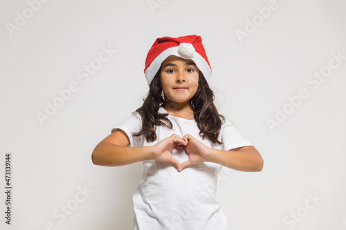 Smiling child girl in christmas santa hat showing heart gesture, congratulating with xmas holiday. Love, care and trust