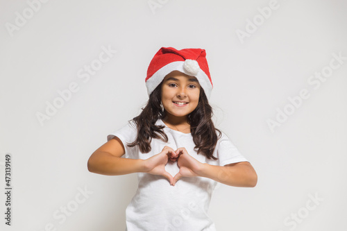 Smiling child girl in christmas santa hat showing heart gesture, congratulating with xmas holiday. Love, care and trust